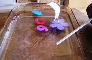 Homemade sailboat and foam shapes floating on water in baking tray. 