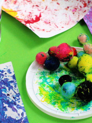 Pompoms in paint sitting on plastic lid surrounded by splatter paint art created by shaken container painting.