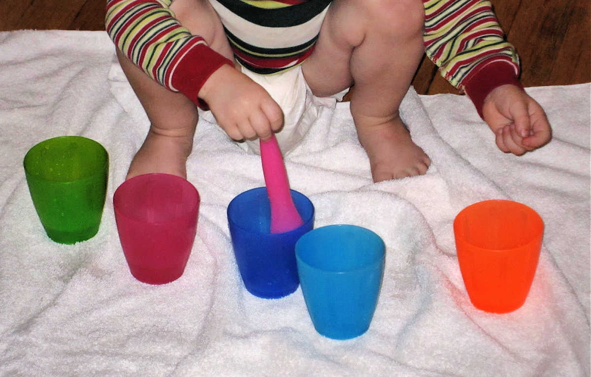 Toddler crouched down on floor, playing with cups and spoon on a white towel.