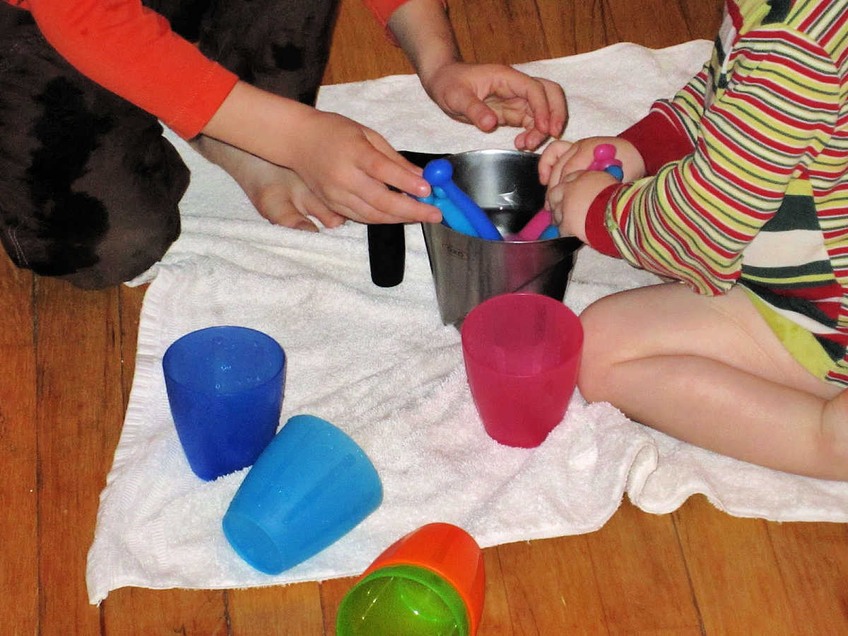 Kids playing with water cups and spoons on a white towel.