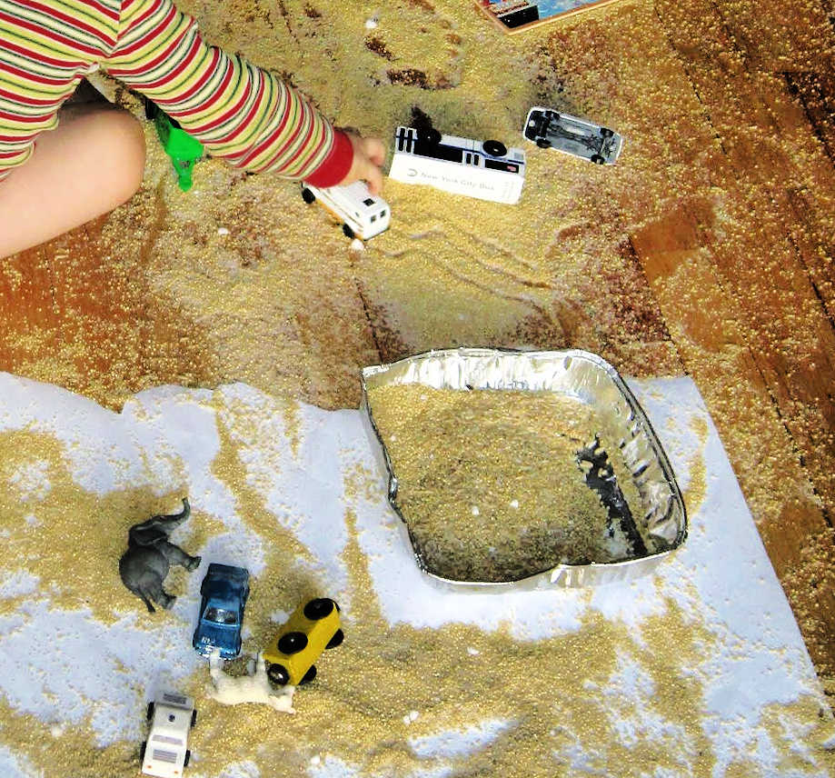 Child playing with cars and making a mess on the floor with a tray of millet