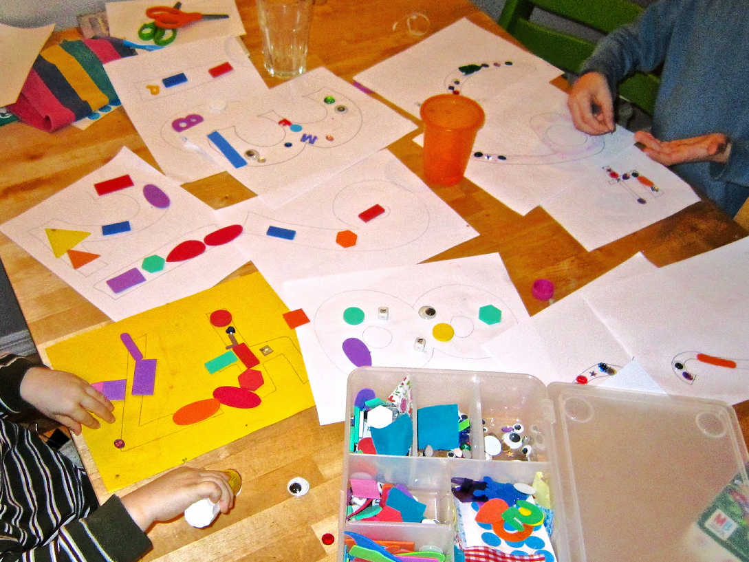 Crafting mess on the table: two children sitting at table using glue to stick collage shapes and materials to paper. Table covered in paper and collage materials.