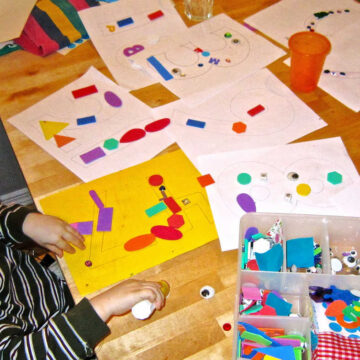 Crafting mess on the table: child sitting at table using glue to stick collage shapes and materials to paper. Table covered in paper and collage materials.