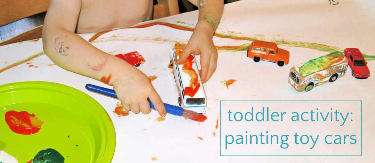 Toddler painting toy bus with orange paint at table covered in paper