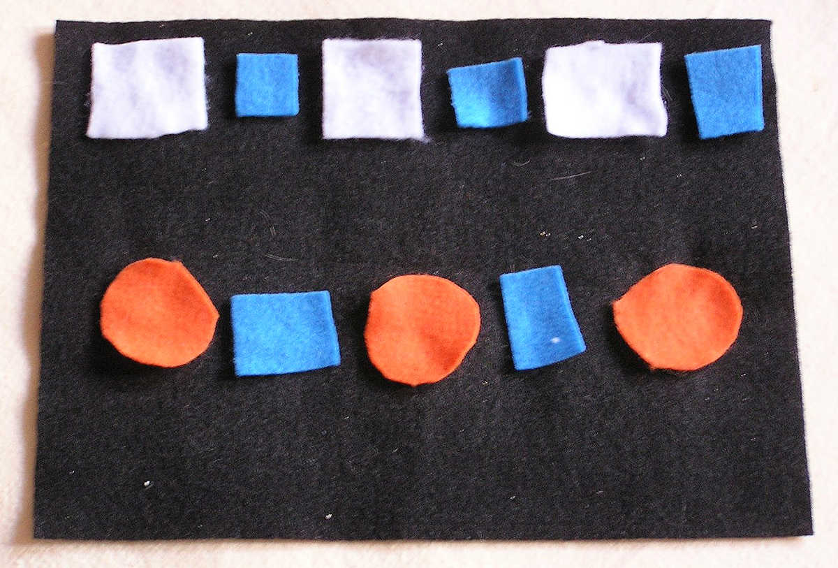 White and blue square and orange circle felt shapes on black background laid out in white square-blue square and orange circle-blue square pattern.