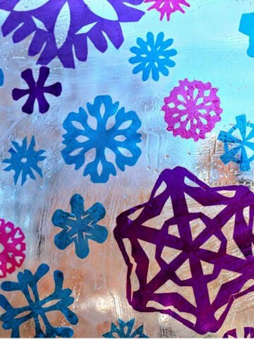 Cellophane snowflakes in blue, pink and purple stuck to window glass, creating a stained glass effect.