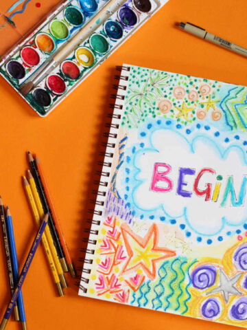 Open page of art journal surrounded by watercolors, pens and pencils.
