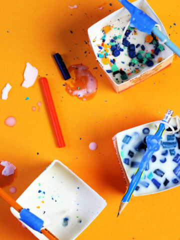 Three milk carton candles in progress, on orange background with scattered wax crayons.
