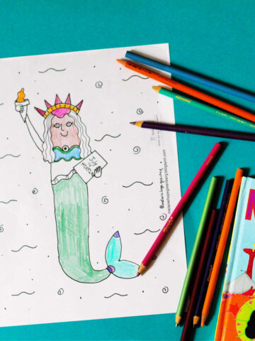 Mermaid with head and arm of statue of liberty coloring page, next to scattered colored pencils and Mermaids on Parade children's picture book.