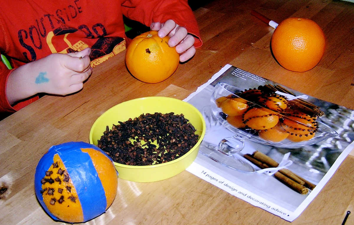 Child poking cloves into orange to make a pomander. Also on table is a dish of cloves, a plain orange, magazine page with photo of pomanders, and a partially completed pomander. 