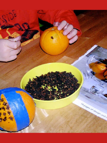Child working on making a pomander, holding an orange, dish of cloves and orange stuck with cloves and bound in blue tape.