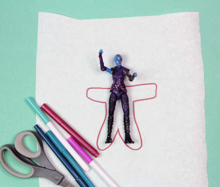 Outline drawn around action figure on white paper, next to pens and scissors.