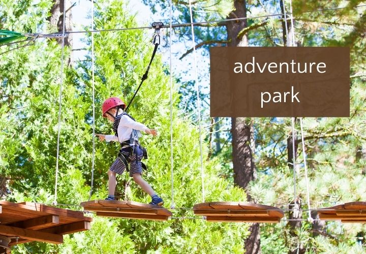 Boy in red helmet and harness walking on treetop obstacle in woods.