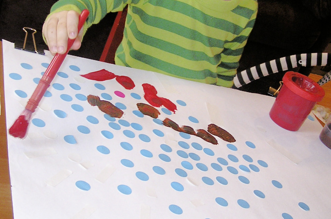 Child making resist art by painting with red paint over blue stickers
