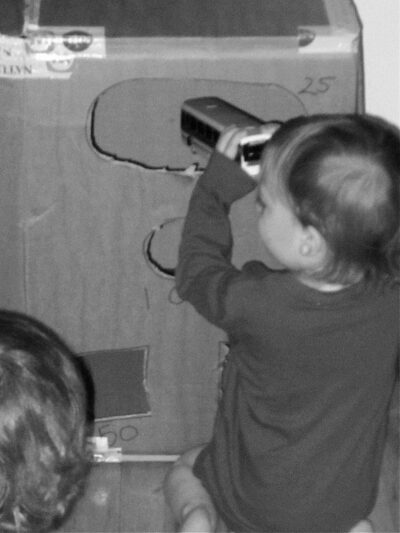 Boy placing toy bus into cut out of diy carnival game make from large cardboard box.