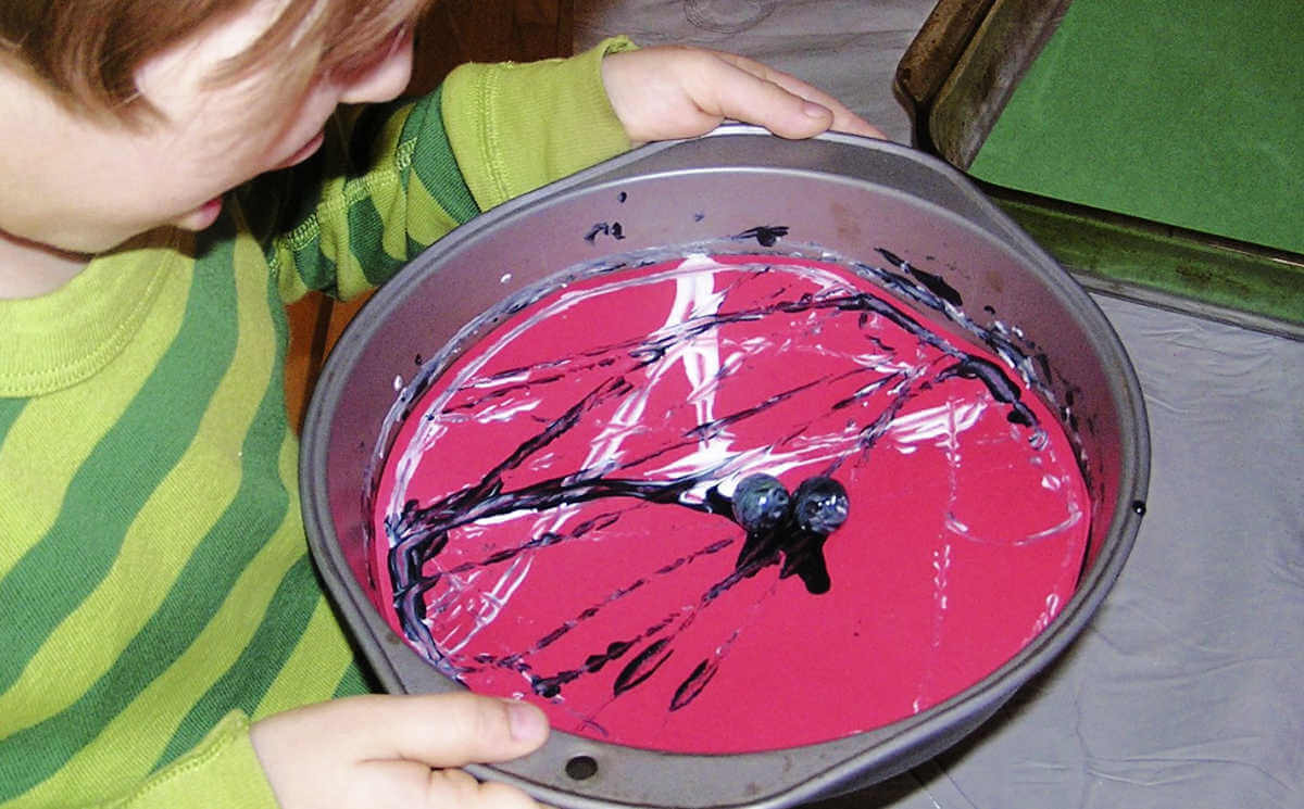 Child holding cake pan with paint covered marbles on red paper