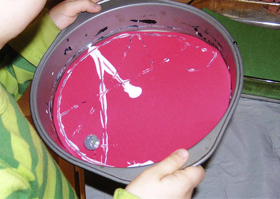 Child holding cake pan with white paint covered marble on red paper