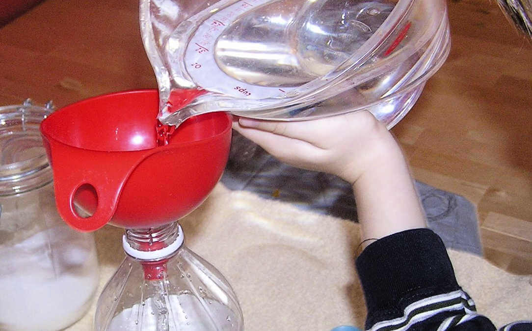 Child's hand pouring water from measuring cup into red funnel resting in bottle neck