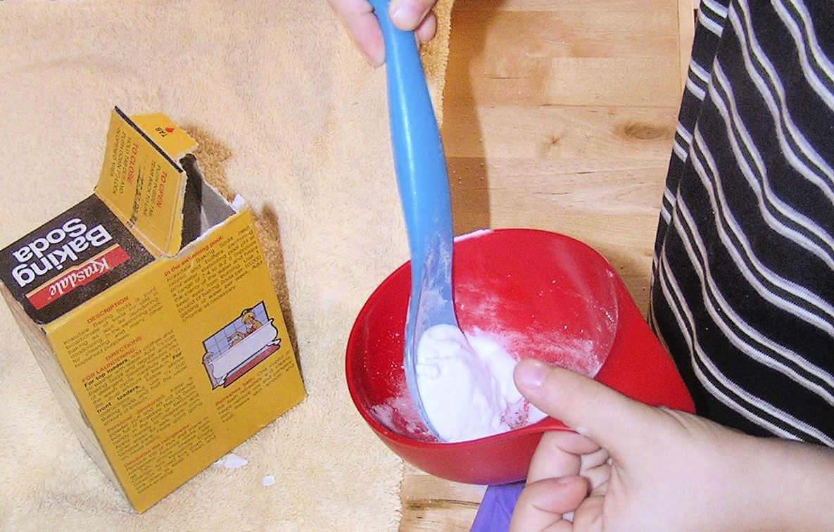 Child putting baking soda into funnel attached to a balloon, next to box of soda on the table