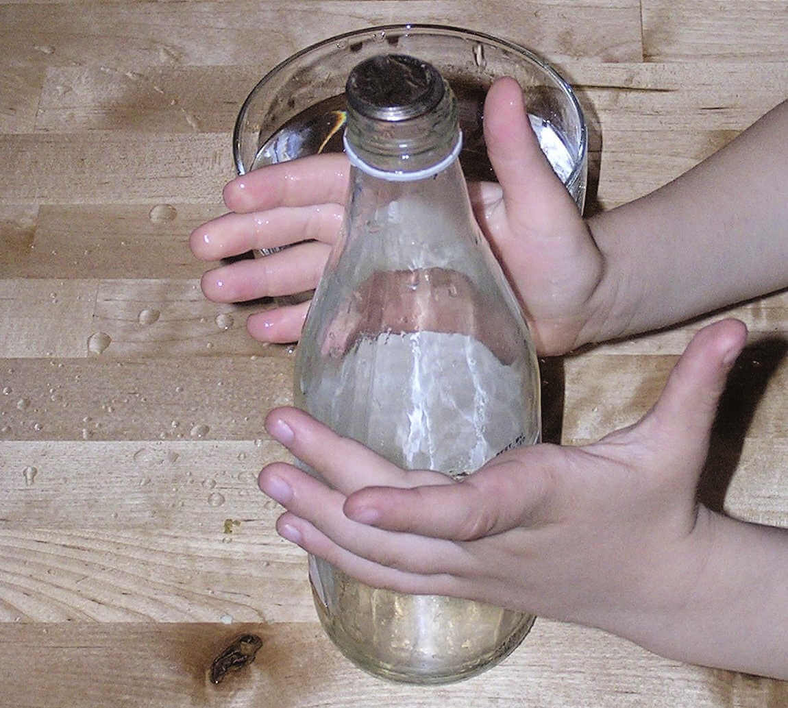 Child hands surrounding glass bottle partially filled with water, with coin on the top in preparation for jumping coin trick