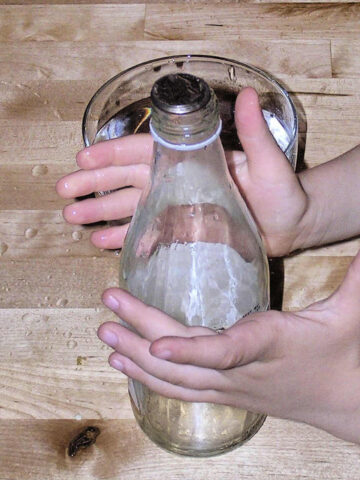 Child hands surrounding glass bottle partially filled with water, with coin on the top for jumping coin trick