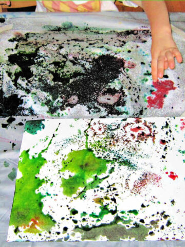 Child working on watercolor and salt art project at messy table covered in painting supplies