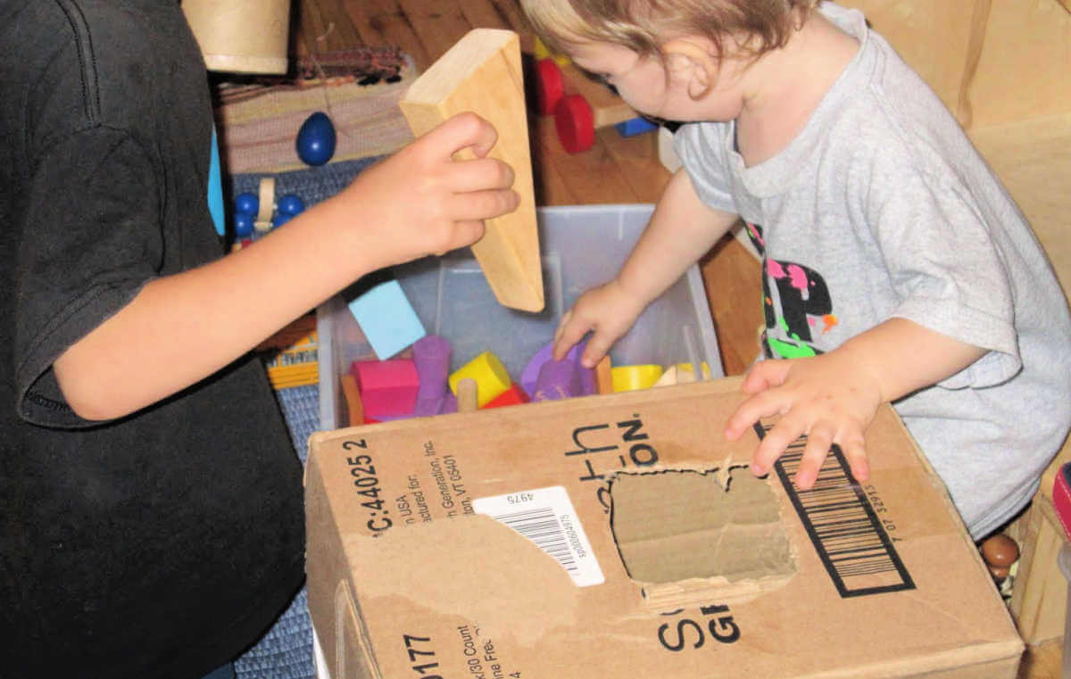 Two children dropping toy blocks into a hole cut in a cardboard box