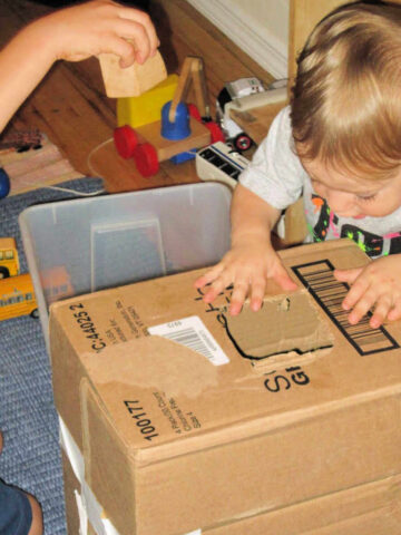 Two children dropping toy blocks into a hole cut in a cardboard box