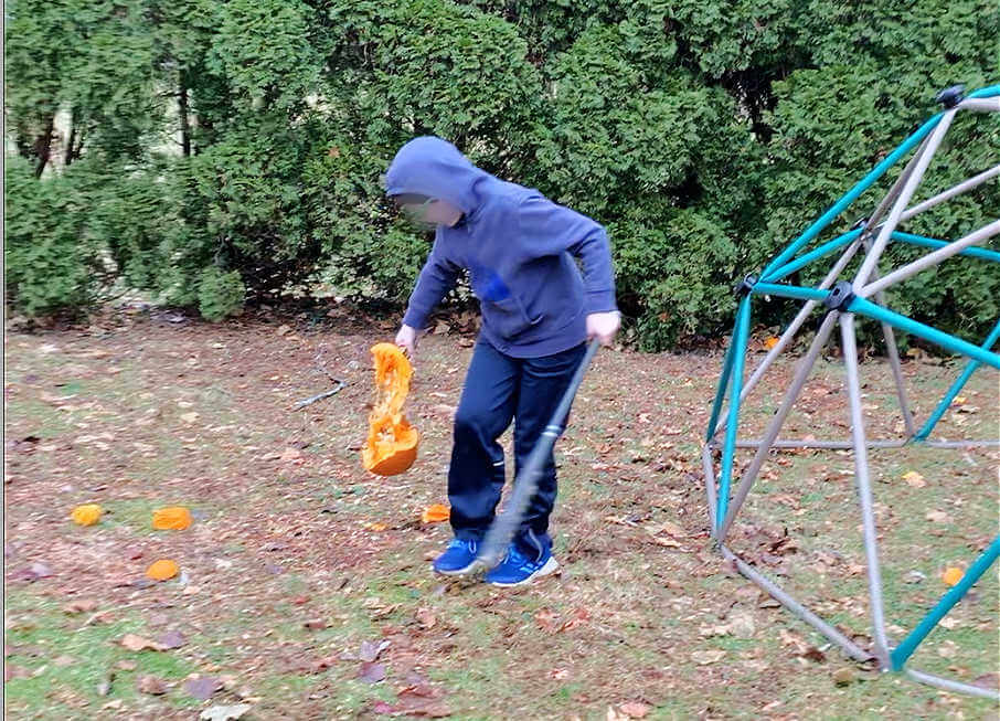 Child picking up broken pumpkin and holding baseball bat.