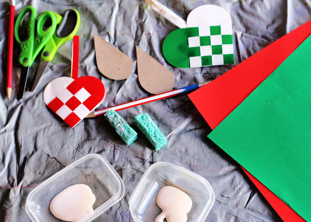 Materials for sponge painted woven heart ornament craft including scissors, paper, paint, sponge and finished woven hearts.