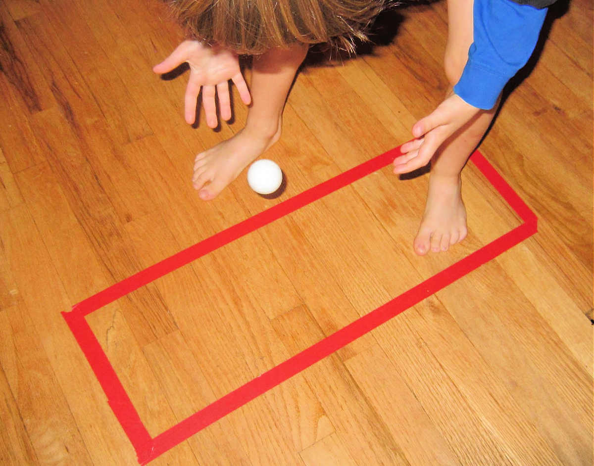 Child dropping ping pong ball onto floor inside of a red tape rectangle.