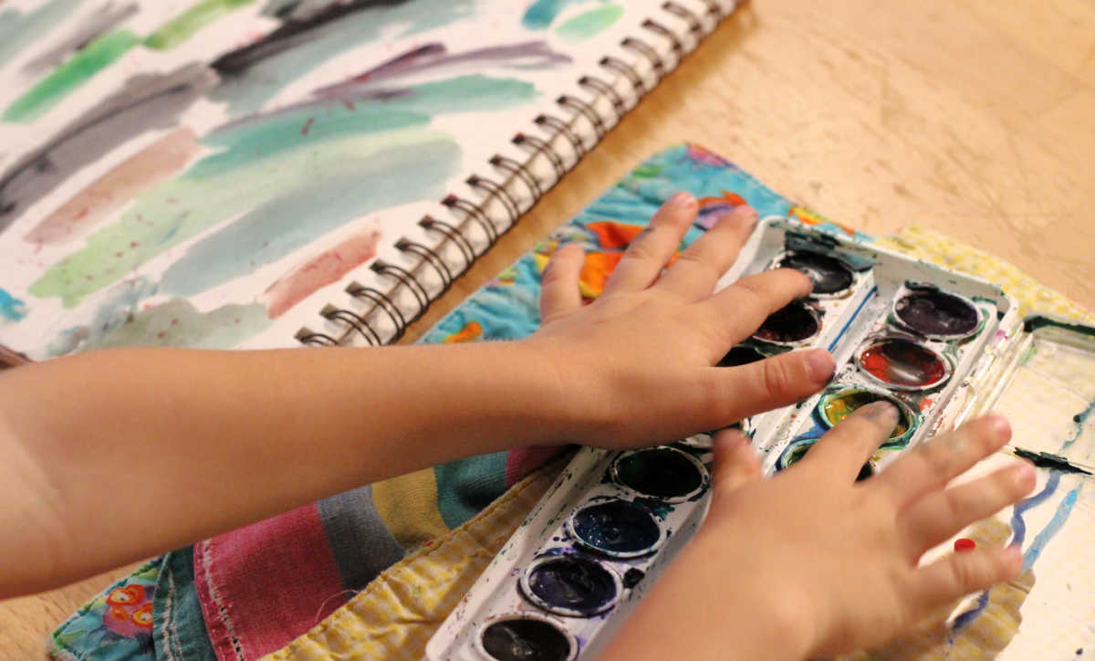 Child's hands putting fingers in watercolor tray with art journal page of paint smears.