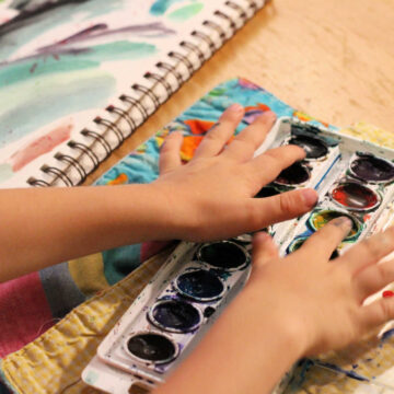 Child's hands putting fingers in watercolor tray with art journal page of paint smears.