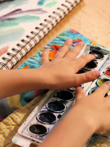 Child's hands putting fingers in watercolor tray with art journal page of paint smears.