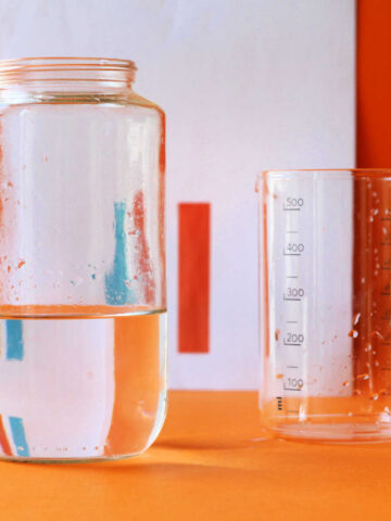 Water refraction experiment materials: jar with water, empty beaker in front of blue and orange bars on a piece of paper.