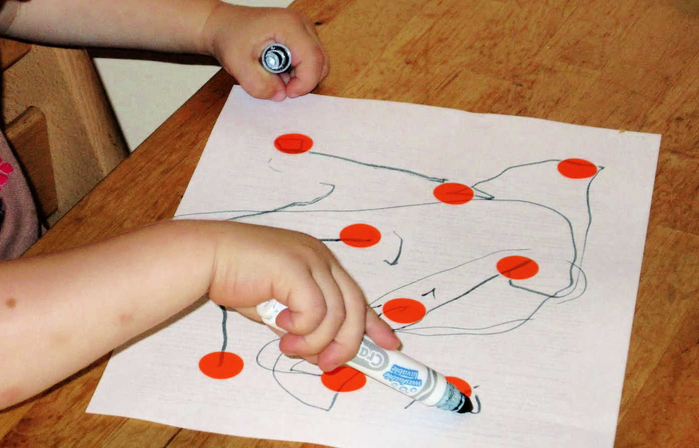 Toddler using large marker to draw on paper covered with orange circle stickers.