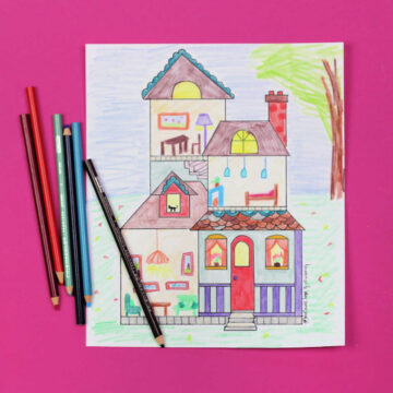 Dollhouse coloring page, colored in and sitting on pink background next to colored pencils.